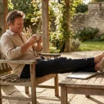 A man relaxes on a wooden patio chair in a sunny garden, drinking coffee.